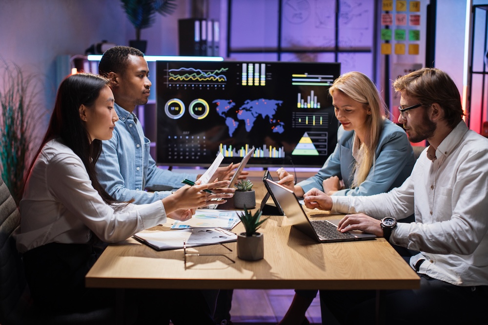 Diverse team of professionals reviewing charts and data on laptops and tablets with world map visualization in background