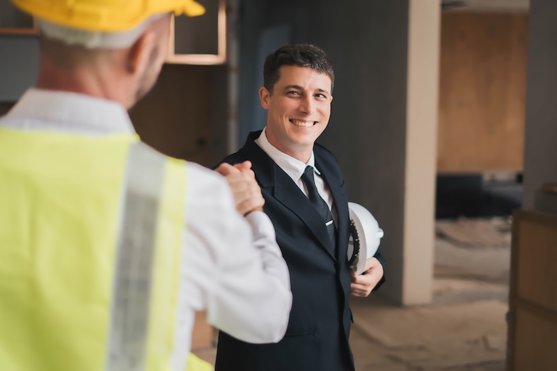 Businessman in suit holding hard hat shaking hands with construction worker at a building site
