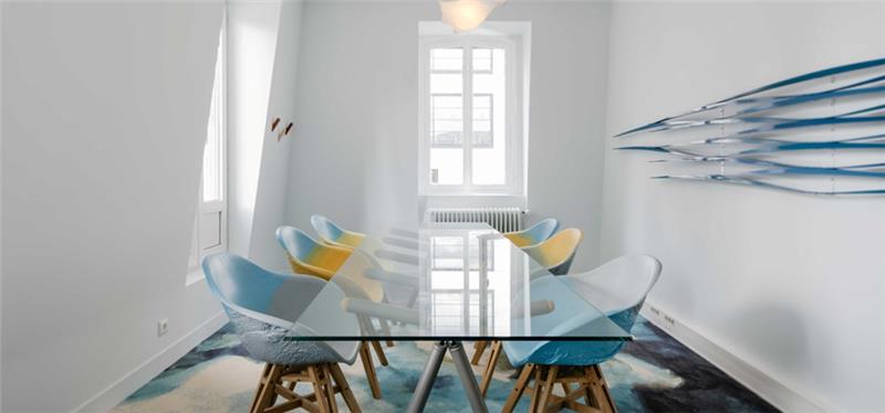 Salle de réunion moderne avec grande table en verre, chaises design upcyclées bleu/jaune et tapis aux tons marbrés — aménagement de bureaux par Lbc.