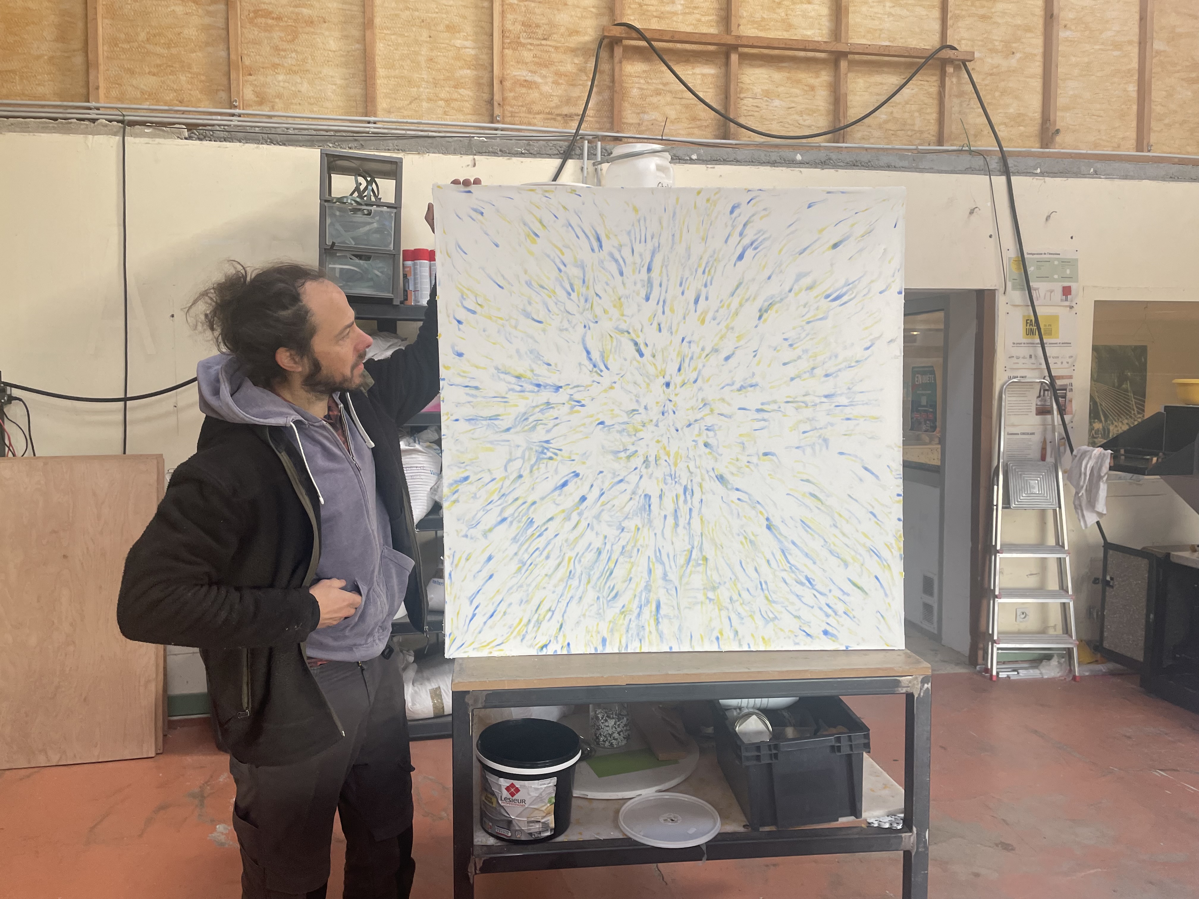 Artiste dans un atelier présentant une grande toile abstraite aux tons blancs, bleus et jaunes, posée sur un établi